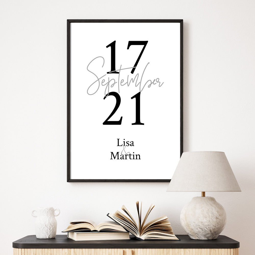 Personalisiertes Poster. Individueller Monat in grauer Schrift mittig, drum rum das Datum. Unten stehen die Vornamen. Bild an der Wand im schwarzen Rahmen.