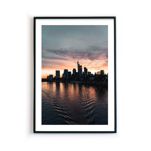 Frankfurt Skyline Poster zum Sonnenuntergang. Kraftvoller Himmel mit Spiegelungen im Main. Bild mit weißen umlaufenden Rand, gerahmt im schwarzen Rahmen.