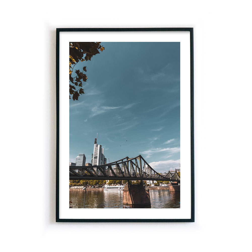 Frankfurt Poster vom Eisernen Steg und der Skyline im Hintergrund. Bild mit weißen umlaufenden Rand. Gerahmt in einem schwarzen Bilderrahmen.