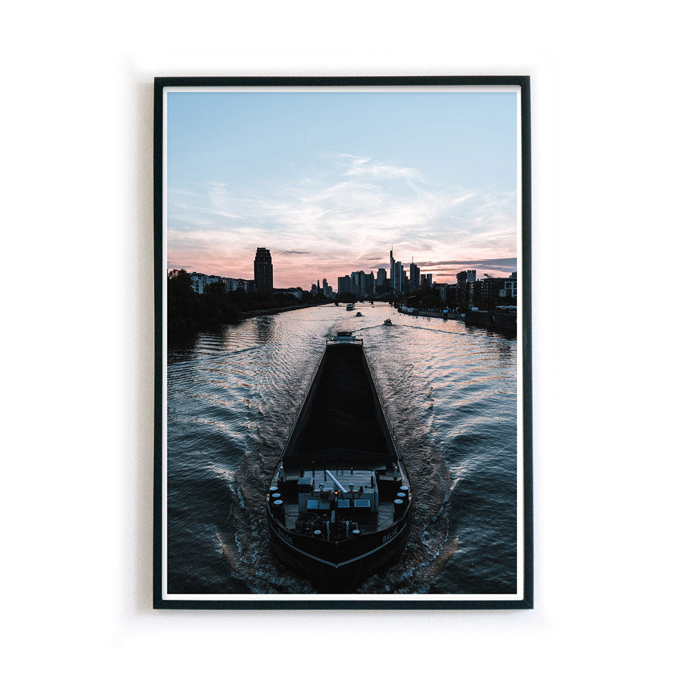 Frankfurt am Main Poster. Boot fährt über den Main, im Hintergrund die Frankfurter Skyline zum Sonnenuntergang. Bild im schwarzen Bilderrahmen.