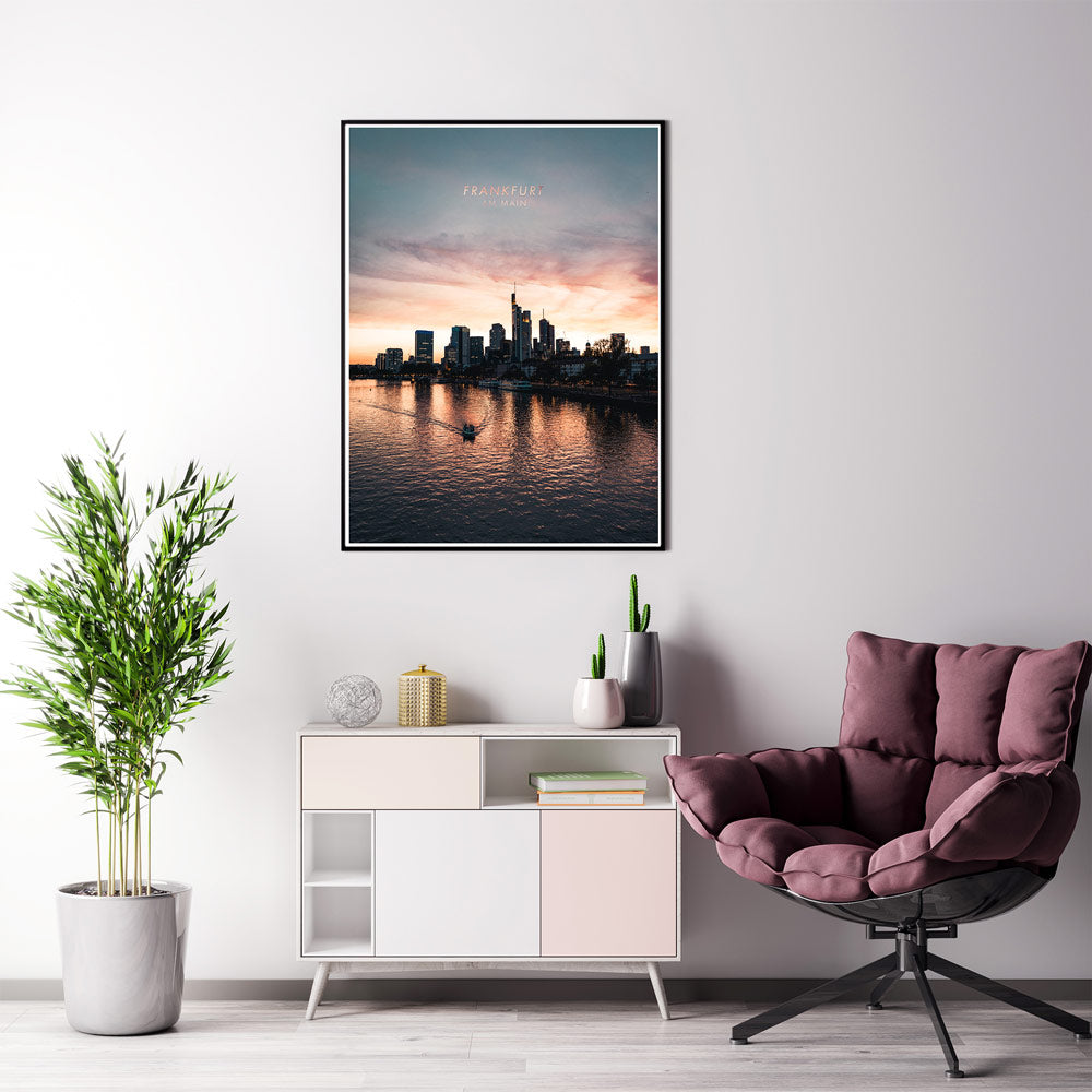 Frankfurt am Main Skyline Poster. Farbenfroher Sonnenuntergang, kleines Boot fährt im Vordergrund über den Main. Bild im schwarzen Bilderrahmen an der Wand im Wohnzimmer.