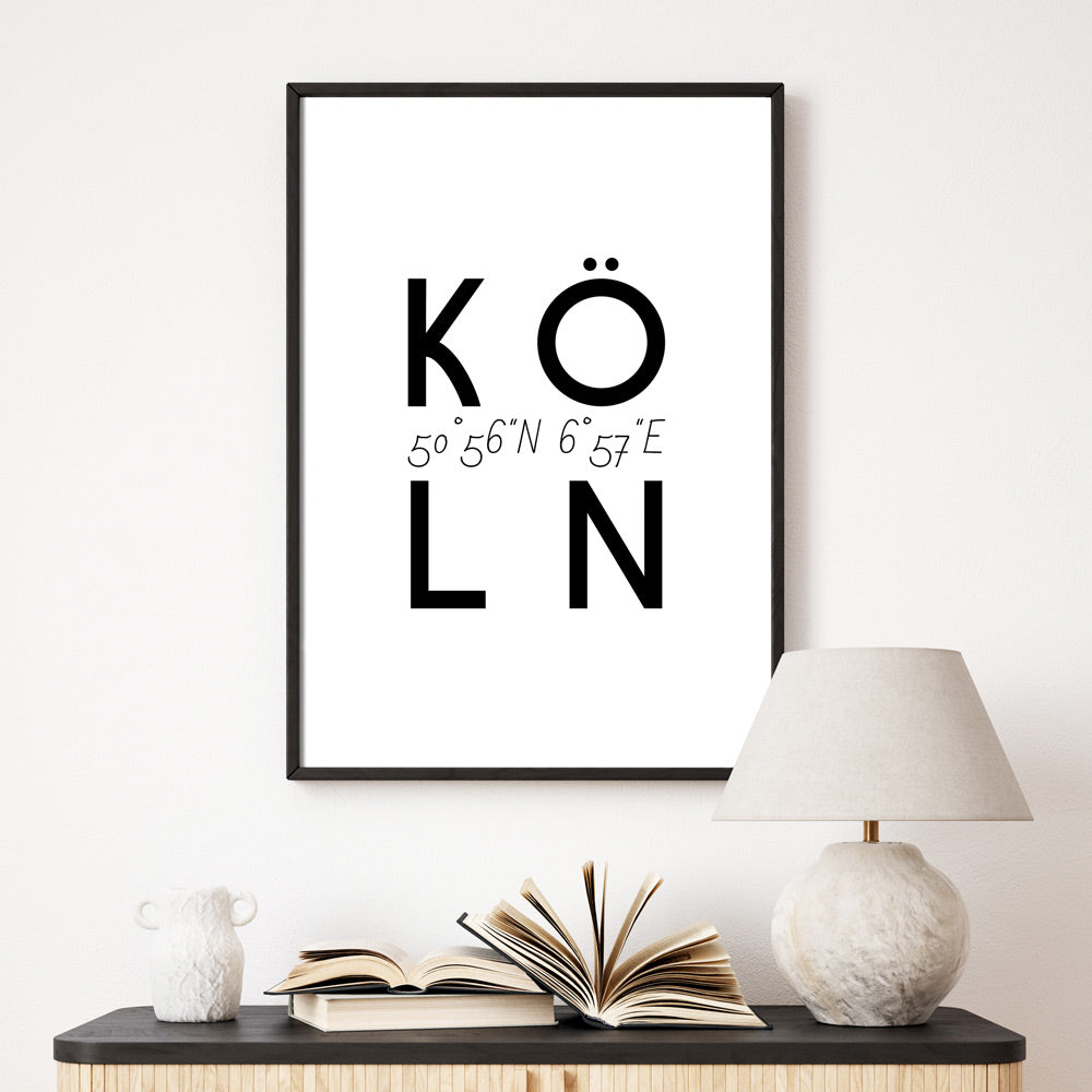 Köln Spruch Bild in schwarz weiß. Köln Schriftzug mit den Köln Koordinaten in der Mitte. Bild im schwarzen Bilderrahmen an der Wand über einer Kommode