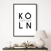 Köln Spruch Bild in schwarz weiß. Köln Schriftzug mit den Köln Koordinaten in der Mitte. Bild im schwarzen Bilderrahmen an der Wand über einer Kommode