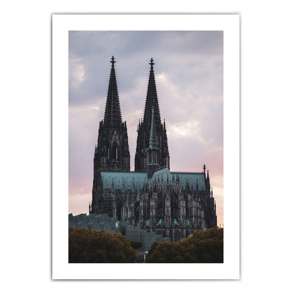 Kölner Dom Poster im Sonnenuntergang mit rötlichem Himmel. Im Vordergrund das Museum Ludwig und Bäume. Bild hat einen weißen umlaufenden Rand.