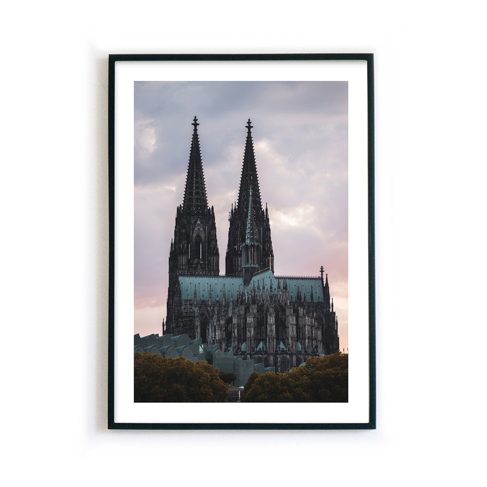 Kölner Dom Poster im Sonnenuntergang mit rötlichem Himmel. Im Vordergrund das Museum Ludwig und Bäume. Bild hat einen weißen Rand und ist gerahmt.