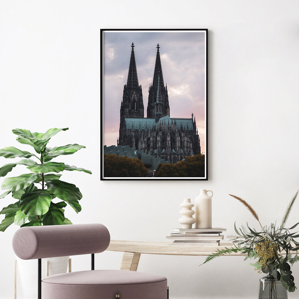 Kölner Dom Poster im Sonnenuntergang mit rötlichem Himmel. Im Vordergrund das Museum Ludwig und Bäume. Fertig gerahmt über einen hellen Tisch und rosa Stuhl.