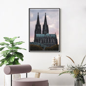 Kölner Dom Poster im Sonnenuntergang mit rötlichem Himmel. Im Vordergrund das Museum Ludwig und Bäume. Fertig gerahmt über einen hellen Tisch und rosa Stuhl.
