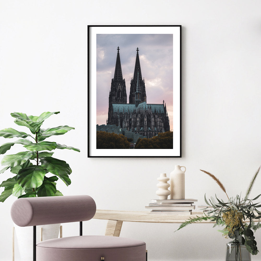 Kölner Dom Poster im Sonnenuntergang mit rötlichem Himmel. Im Vordergrund das Museum Ludwig und Bäume. Fertig gerahmt mit weißen Rand aufgehangen im Wohnzimmer.