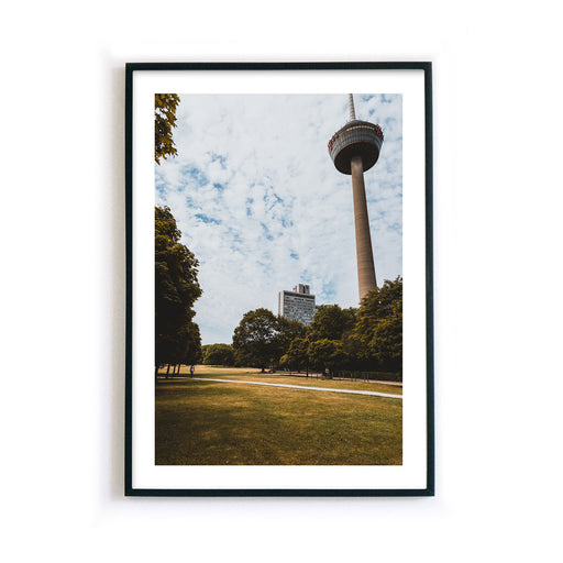 Retro Köln Poster vom Fernsehturm im grünen Gürtel. Grüne Wiese und Bäume im Vordergrund bei blauem bewölkten Himmel. Bild mit weißen Rand im Rahmen.