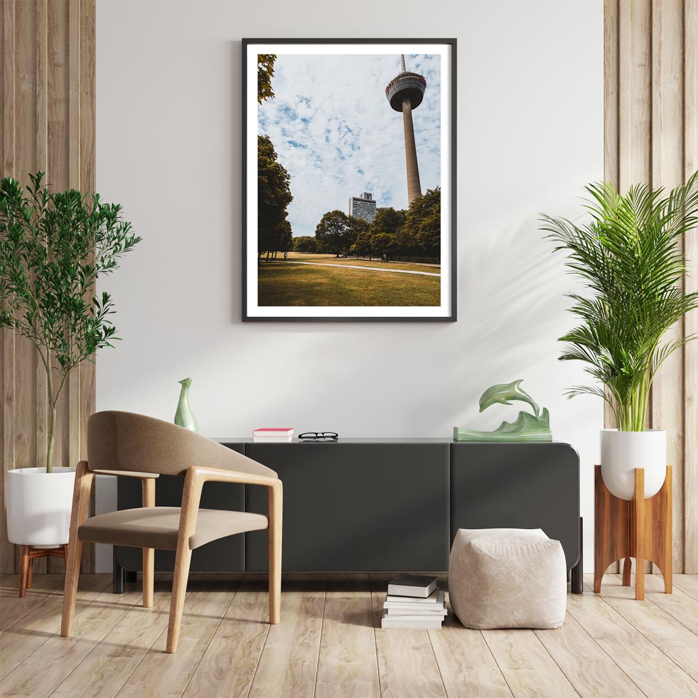Retro Köln Poster vom Fernsehturm im grünen Gürtel. Grüne Wiese und Bäume im Vordergrund bei blauem bewölkten Himmel. Bild mit weißen Rand im Rahmen an der Wohnzimmer-Wand.