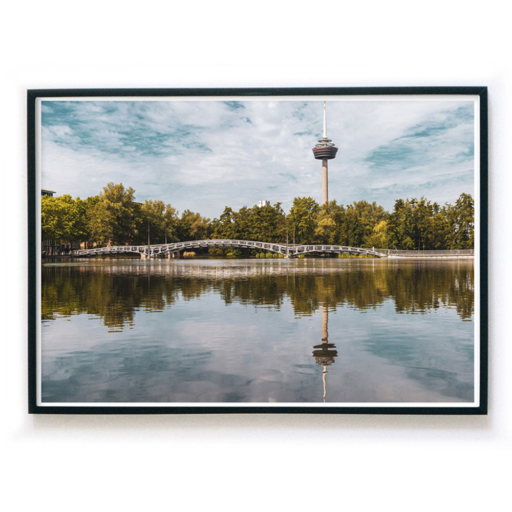 Querformates Köln Bild im schwarzen Bilderrahmen. Motiv ist der Fernsehturm, eine Brücke und Bäume, die sich im See spiegeln. Sommertag mit blauem Himmel.