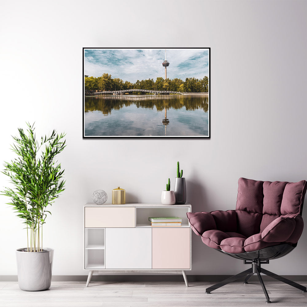Köln Bild im Querformat. Motiv ist der Fernsehturm, eine Brücke und Bäume, die sich im See spiegeln. Im Rahmen über einer Kommode und Sessel an der Wand.