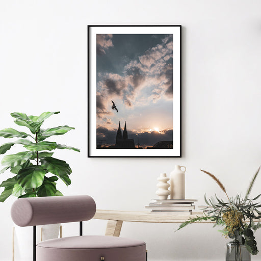 Köln Poster vom Sonnenuntergang über dem Kölner Dom. Über dem Dom fliegt eine Möwe. Kraftvoller blauer Himmel mit Wolken und Sonnenstrahlen. Aufgehangen im schwarzen Rahmen an der Wand im Wohnzimmer. Mit weißen Rand um das Poster.